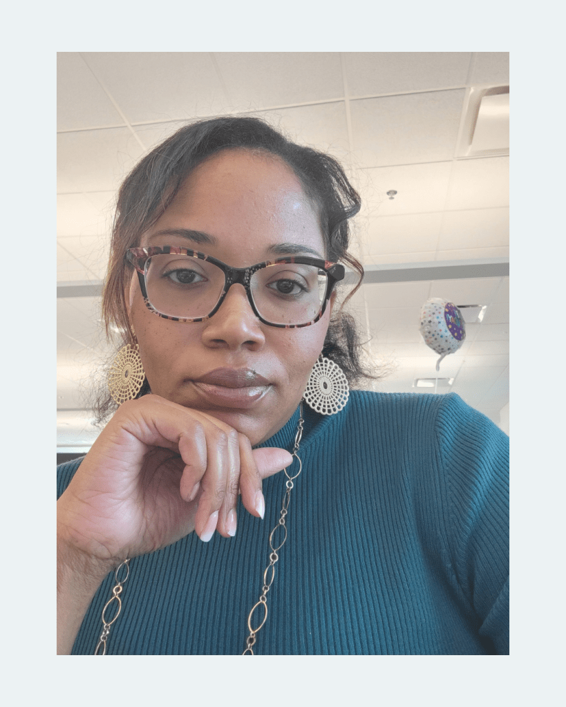 Photo of Anastasia sitting at her desk, chin resting on her hand. Anastasia's hair is pulled back, with her bangs swept to the side. She is wearing pink and black tortoiseshell glasses, a blue turtleneck, gold disc earrings, and a gold draped chain.